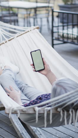 Video-Vertical-De-Una-Mujer-Caucásica-Mayor-Usando-Un-Teléfono-Inteligente-Con-Espacio-Para-Copiar-En-Hamaca,-Cámara-Lenta