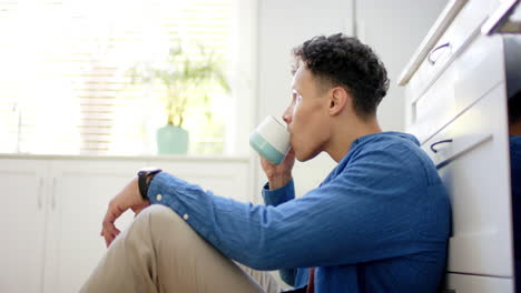 Hombre-Birracial-Tomando-Café-Sentado-En-El-Suelo-En-La-Cocina-De-Casa,-En-Cámara-Lenta