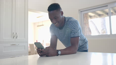 Happy-african-american-man-leaning-on-counter-and-using-smartphone-in-sunny-kitchen,-slow-motion