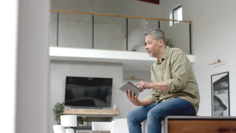 Happy-senior-biracial-man-sitting-on-table-using-tablet-at-home,-copy-space,-slow-motion