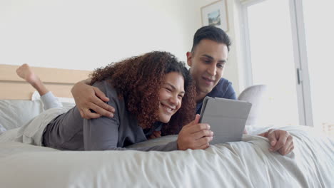 Happy-diverse-gay-male-couple-lying-on-bed-using-tablet-and-embracing-in-the-morning,-slow-motion