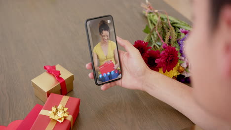 Mujer-Birracial-Sosteniendo-Un-Teléfono-Inteligente-Con-Una-Mujer-Hablando-En-La-Pantalla-Con-Regalos-En-El-Escritorio