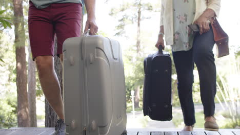 Diverse-senior-couple-walking-with-luggage-to-a-house-in-sunny-outdoors