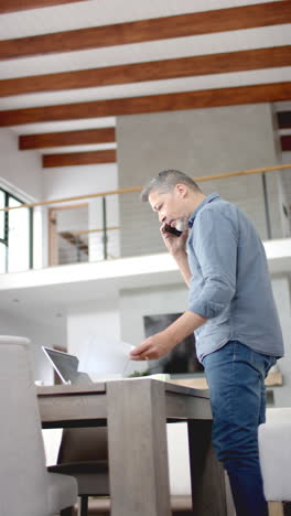 Vertical-video-of-senior-biracial-man-talking-on-smartphone-standing-at-desk-at-home,-slow-motion