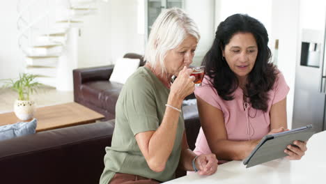 Happy-diverse-senior-women-using-tablet-and-drinking-tea-in-sunny-kitchen,-slow-motion