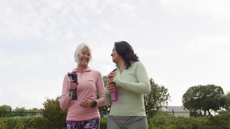 Two-diverse-senior-women-walking-with-water-bottles-discussing-on-sunny-day,-slow-motion,-copy-space