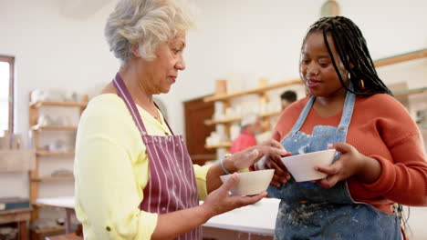 Happy-diverse-female-potters-holding-bowls-and-discussing-in-pottery-studio,-slow-motion