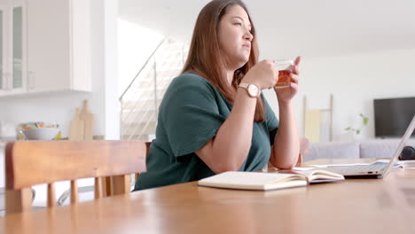 Mujer-Birracial-Reflexiva-De-Talla-Grande-Bebiendo-Té-Y-Usando-Una-Computadora-Portátil,-Trabajando-Desde-Casa,-Cámara-Lenta