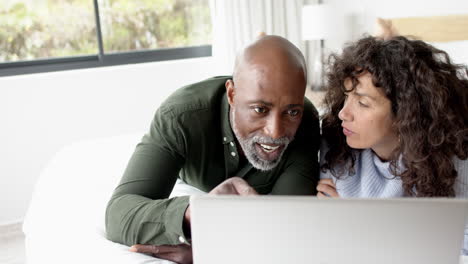 Happy-mature-diverse-couple-lying-on-bed-using-laptop-at-home,-slow-motion