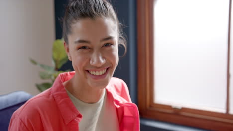 Portrait-of-happy-biracial-teenage-girl-sitting-by-window-at-home-smiling,-copy-space,-slow-motion