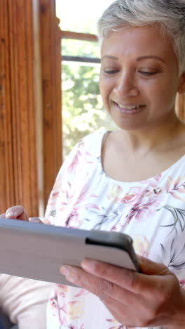 Video-Vertical-De-Una-Feliz-Mujer-Birracial-Mayor-Usando-Una-Tableta-En-Casa,-Cámara-Lenta