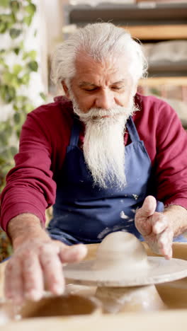 Focused-biracial-male-potter-with-long-beard-using-potter's-wheel-in-pottery-studio,-slow-motion