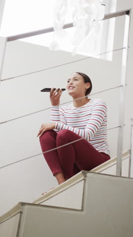 Vertical-video-of-happy-biracial-woman-sitting-on-stairs-talking-on-smartphone,-slow-motion