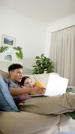 Vertical-video-of-happy-diverse-couple-sitting-on-sofa-using-laptop-at-home,-in-slow-motion