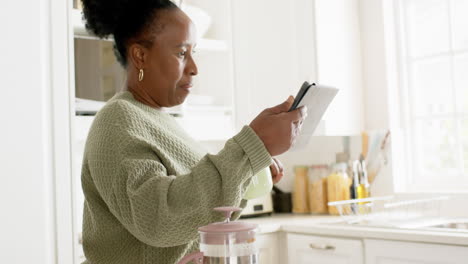 Feliz-Mujer-Mayor-Afroamericana-Tomando-Café,-Usando-Una-Tableta-En-Una-Cocina-Soleada,-Cámara-Lenta