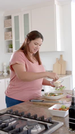 Vertical-video-of-plus-size-biracial-woman-preparing-healthy-salad-in-kitchen,-slow-motion