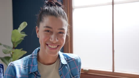 Portrait-of-happy-biracial-teenage-girl-sitting-by-window-at-home-smiling,-copy-space,-slow-motion