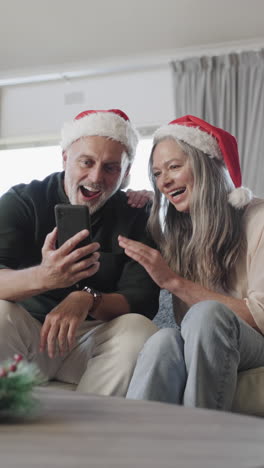 Vertical-video-of-happy-senior-caucasian-couple-wearing-santa-hats-using-smartphone,-slow-motion