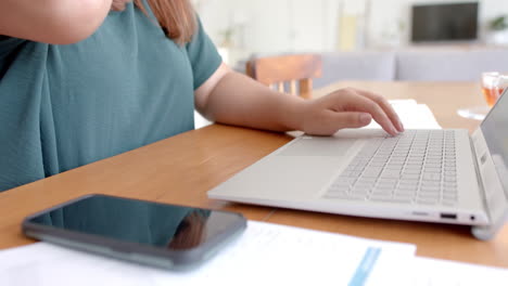 Midsection-of-plus-size-biracial-woman-using-laptop-at-table,-working-from-home,-slow-motion