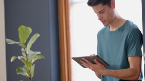 Focused-biracial-man-using-tablet-standing-in-sunny-living-room,-copy-space,-slow-motion