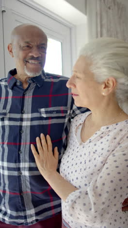 Video-Vertical-De-Una-Feliz-Pareja-Birracial-Mayor-Abrazándose-En-La-Ventana-De-Casa,-Cámara-Lenta