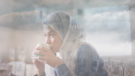 Animation-of-cityscape-over-biracial-woman-in-hijab-drinking-tea