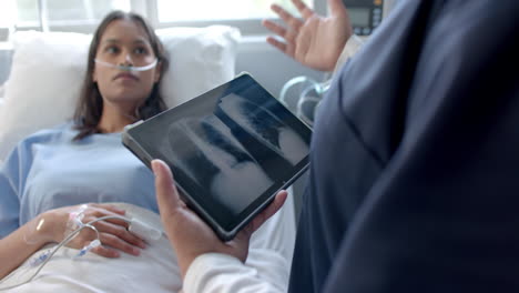 Diverse-female-patient-in-hospital-bed-and-female-doctor-using-tablet-talking,-slow-motion