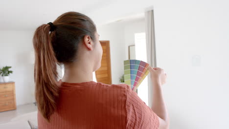 Mujer-Birracial-Pensativa-De-Talla-Grande-Mirando-Cartas-De-Colores-De-Pintura-En-Una-Sala-De-Estar-Soleada,-Cámara-Lenta