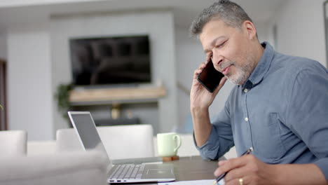 Hombre-Birracial-Mayor-Hablando-Por-Teléfono-Inteligente-Y-Usando-Una-Computadora-Portátil-En-La-Mesa,-Trabajando-Desde-Casa,-Cámara-Lenta