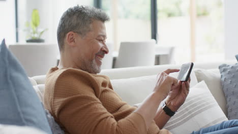 Happy-senior-biracial-man-using-smartphone-on-couch,-laughing-in-sunny-living-room,-slow-motion