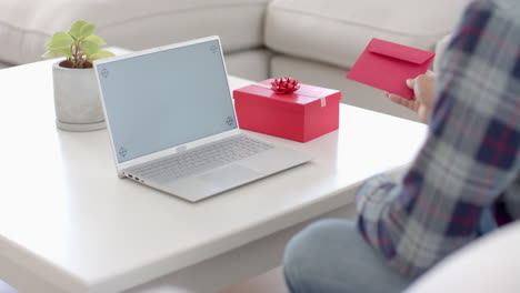 Biracial-man-with-card-and-gift-having-laptop-video-call-with-copy-space-screen-at-home,-slow-motion