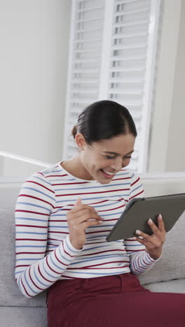 Vertical-video-of-happy-biracial-woman-sitting-on-sofa-using-tablet,-slow-motion