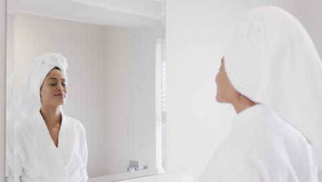 Happy-biracial-woman-wearing-towel-on-head-looking-in-mirror-in-bathroom,-slow-motion