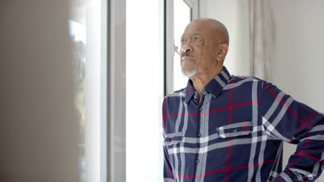 Thoughtful-senior-biracial-man-looking-through-window-at-home,-slow-motion