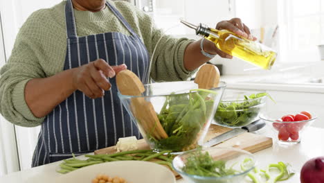 Feliz-Mujer-Mayor-Afroamericana-Mezclando-Ensalada-En-Una-Cocina-Soleada,-Cámara-Lenta