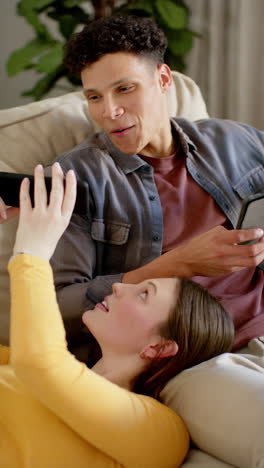 Vertical-video-of-happy-diverse-couple-sitting-on-sofa-using-tablet-and-smartphone,-in-slow-motion