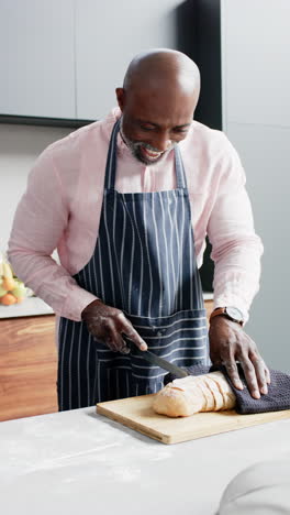 Vertical-video-of-happy-mature-african-american-man-slicing-fresh-bread-in-kitchen,-slow-motion