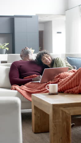 Vertical-video-of-diverse-mature-couple-using-laptop-laughing-on-couch,-copy-space,-slow-motion