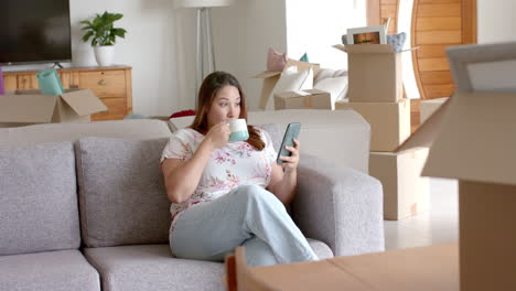 Mujer-Birracial-De-Talla-Grande-En-Un-Sofá-Usando-Un-Teléfono-Inteligente-Tomando-Café-En-Un-Nuevo-Hogar,-Espacio-Para-Copiar,-Cámara-Lenta