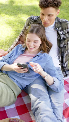 Vertical-video-of-diverse-couple-having-picnic-and-using-smartphone-in-sunny-garden,-in-slow-motion