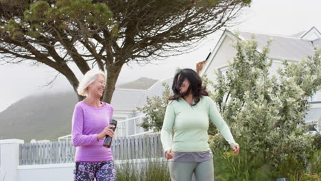 Two-diverse-senior-women-walking-water-bottles-talking-on-sunny-day,-slow-motion