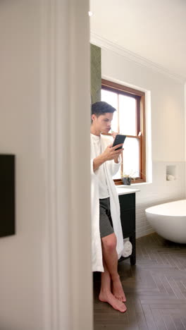 Vertical-video-of-biracial-man-brushing-teeth-using-smartphone-in-bathroom,-copy-space,-slow-motion