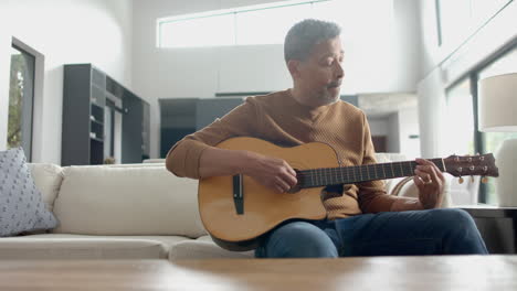 Hombre-Birracial-Senior-Enfocado-Sentado-En-El-Sofá-Tocando-La-Guitarra-En-Casa,-Copiando-Espacio,-Cámara-Lenta