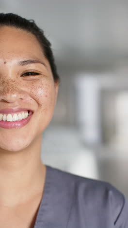 Vertical-video-of-portrait-of-happy-asian-female-doctor-wearing-scrubs-in-hospital,-slow-motion