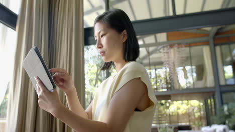 Happy-asian-woman-standing-and-using-tablet-in-sunny-living-room,-slow-motion