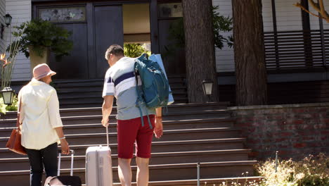 Happy-diverse-senior-couple-walking-with-luggage-to-a-house-in-sunny-outdoors