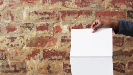 Video-of-hand-of-african-american-man-holding-book-with-blank-pages,-copy-space-over-brick-wall