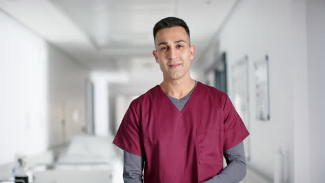 Portrait-of-happy-biracial-male-doctor-wearing-scrubs-in-hospital,-slow-motion