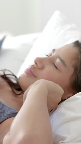 Vertical-video-of-biracial-teenage-girl-lying-in-bed-stretching-on-sunny-morning-slow-motion