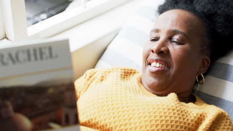 Happy-african-american-senior-woman-lying,-reading-book-and-smiling-in-sunny-room,-slow-motion